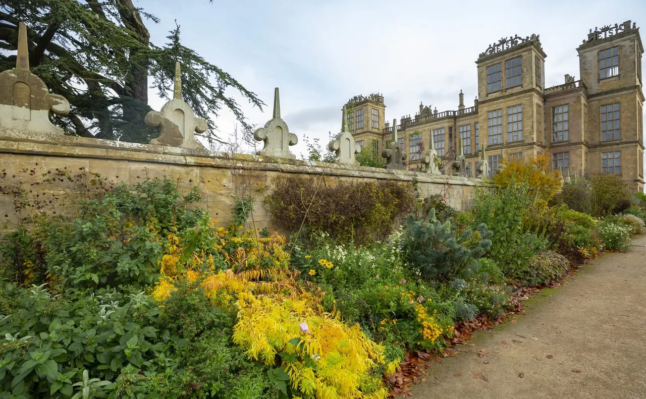 A walled garden path with autumn-flowering plants and shrubs alongside a stone wall topped with ornamental finials, with a large Elizabethan mansion visible beyond.