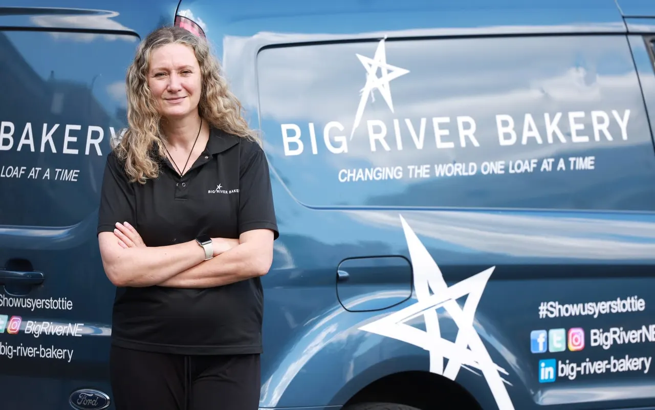 Woman in black Big River Bakery uniform standing with arms crossed in front of a blue bakery van against a blue sky.