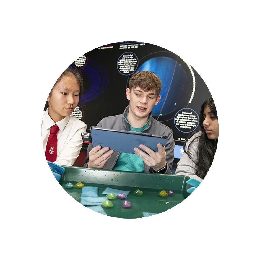 Three young people look at a tablet whilst interacting with a hands-on science exhibit featuring coloured gems on a green table.