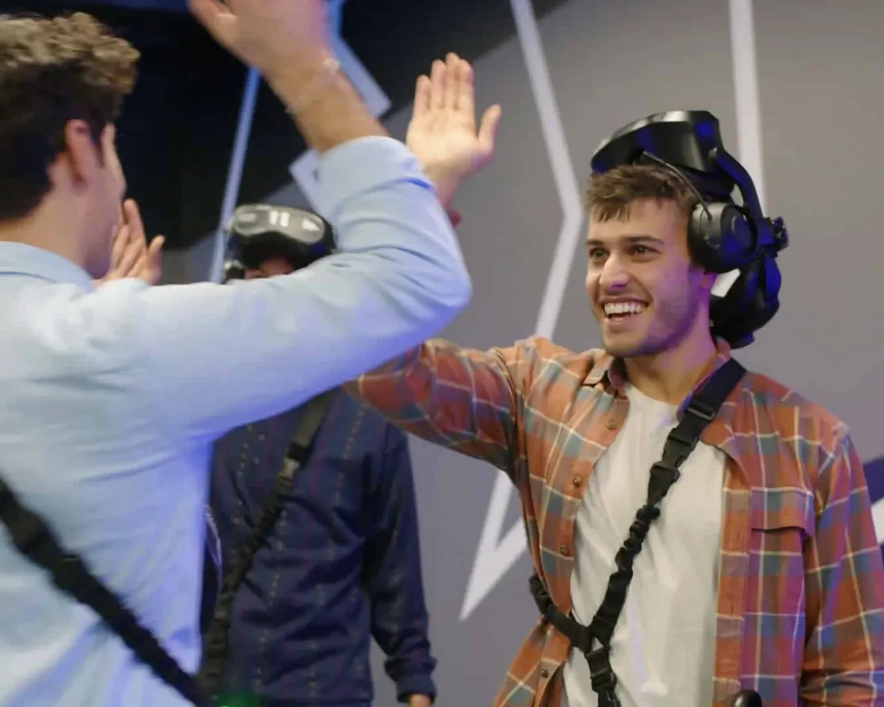 Two young men high-five whilst wearing VR headsets and handheld controllers in an indoor virtual reality gaming space.