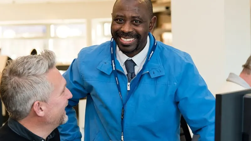 Staff member in blue jacket smiling whilst greeting a visitor in a library reception area.