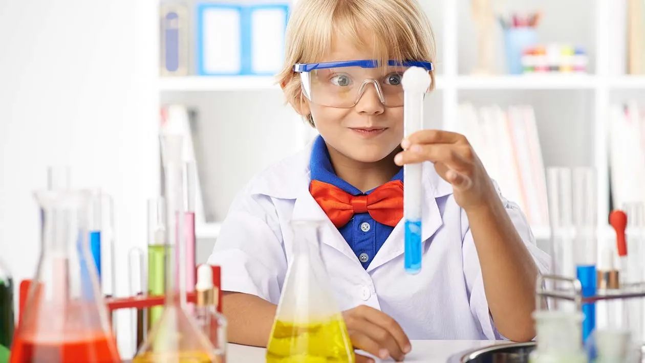 Child wearing safety goggles and lab coat conducts a science experiment with coloured liquids in test tubes.