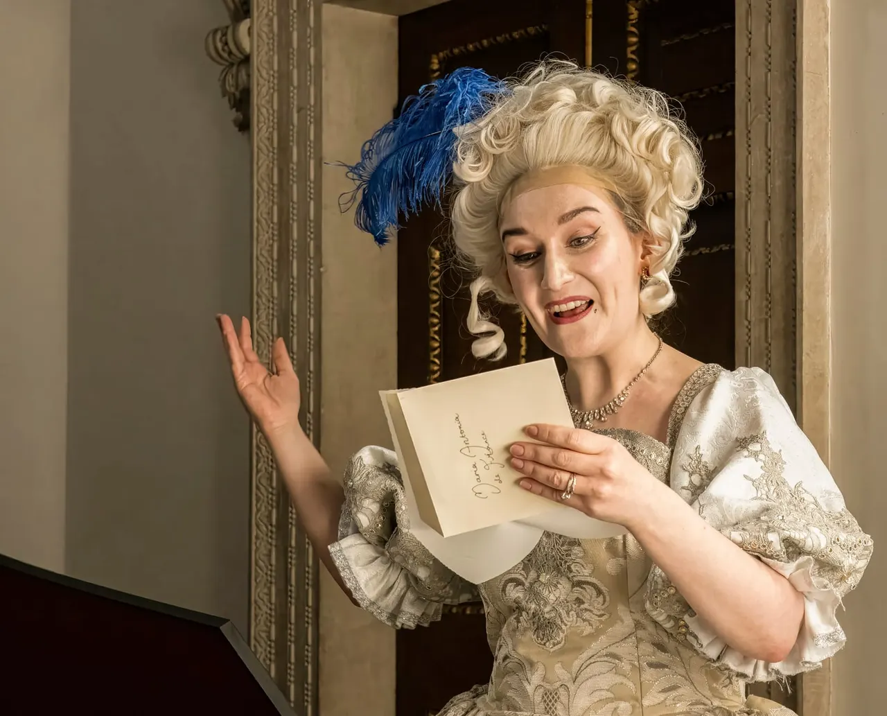 A performer in 18th-century costume with a blue feathered headdress reads from a letter in an ornate period room.
