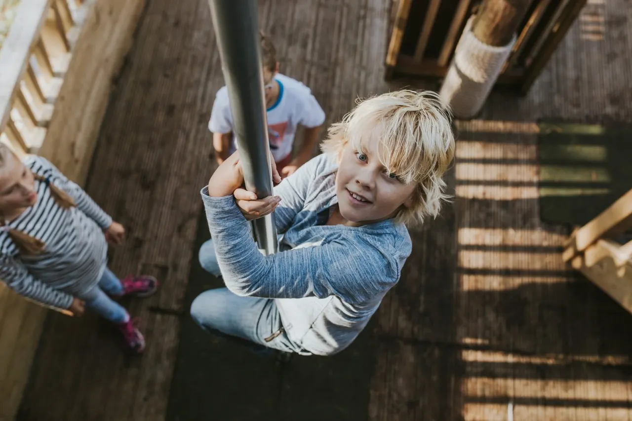 Young child swinging on a rope in a wooden play structure with other children nearby.