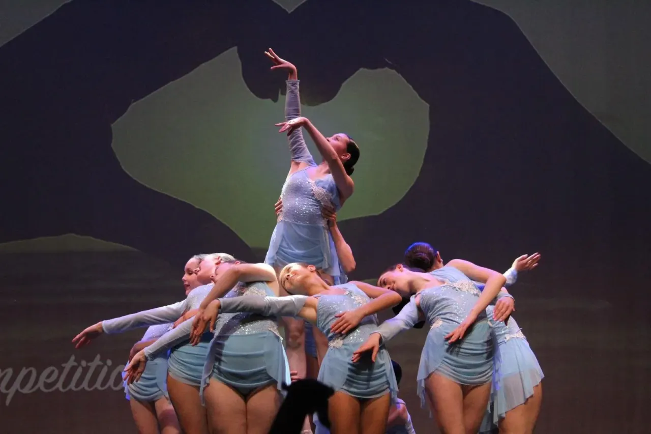 Acrobatic dance performance with multiple dancers in light blue costumes, one balanced on others' shoulders with arm raised.