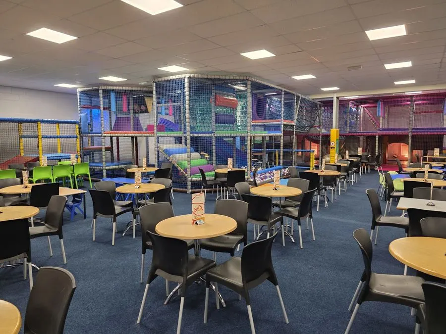 Indoor play centre with dining area featuring round tables, chairs, and colourful multi-level soft play structures behind netting.