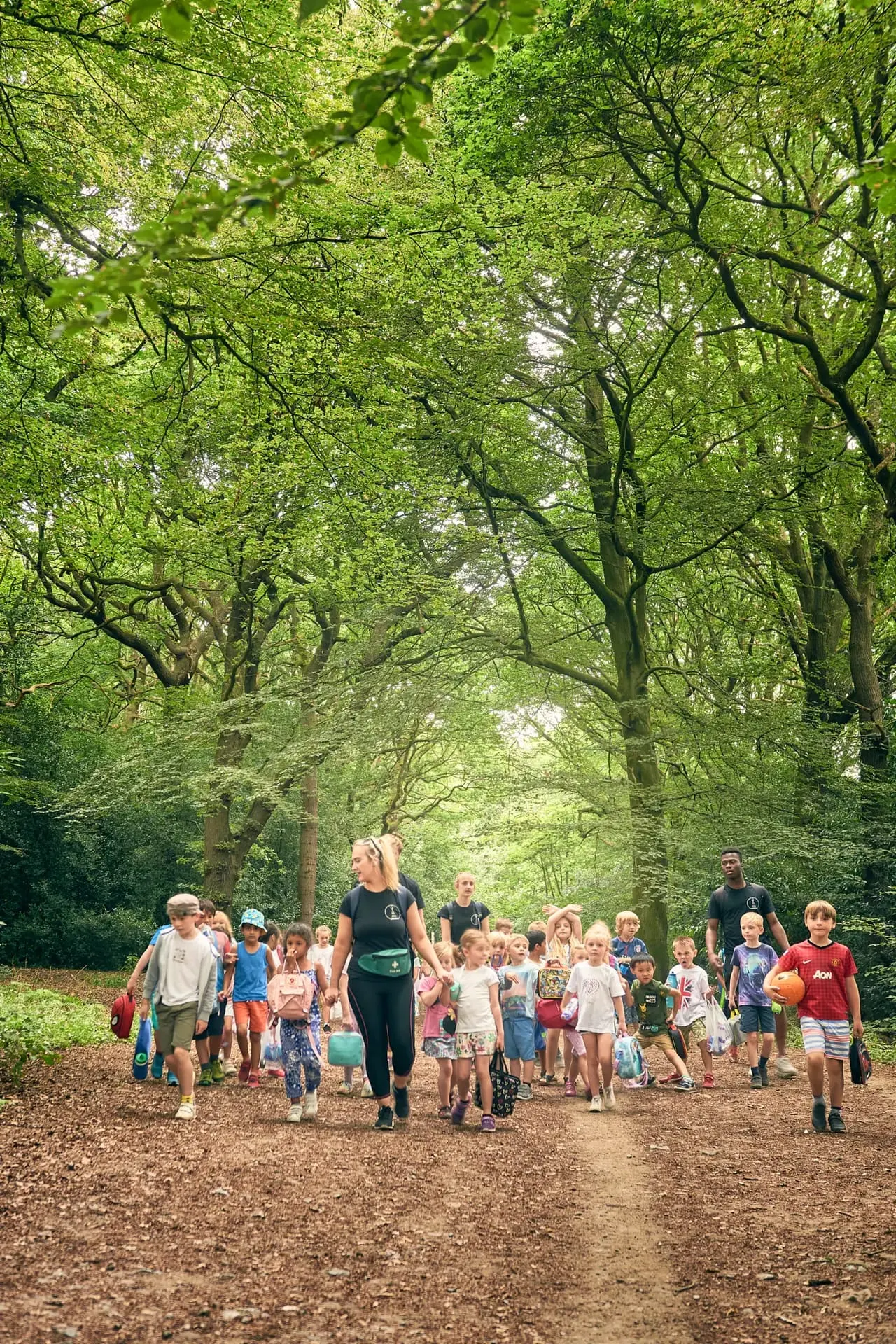 Children and adults walking along a tree-lined woodland path during a guided group activity.
