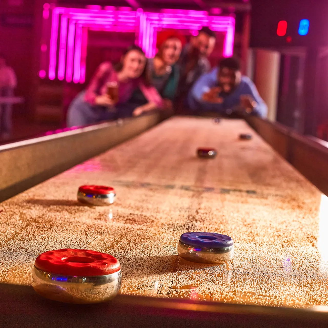 Group of people playing shuffleboard under neon pink lighting in an entertainment venue.