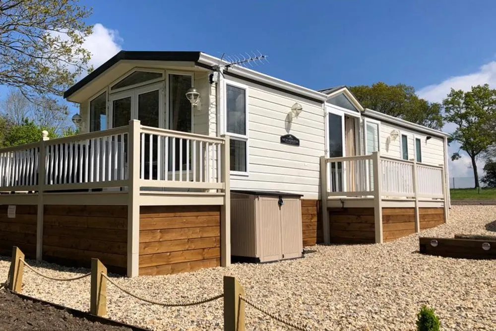 Modern holiday lodge with decking and white railings on a gravel site, surrounded by trees under blue sky.