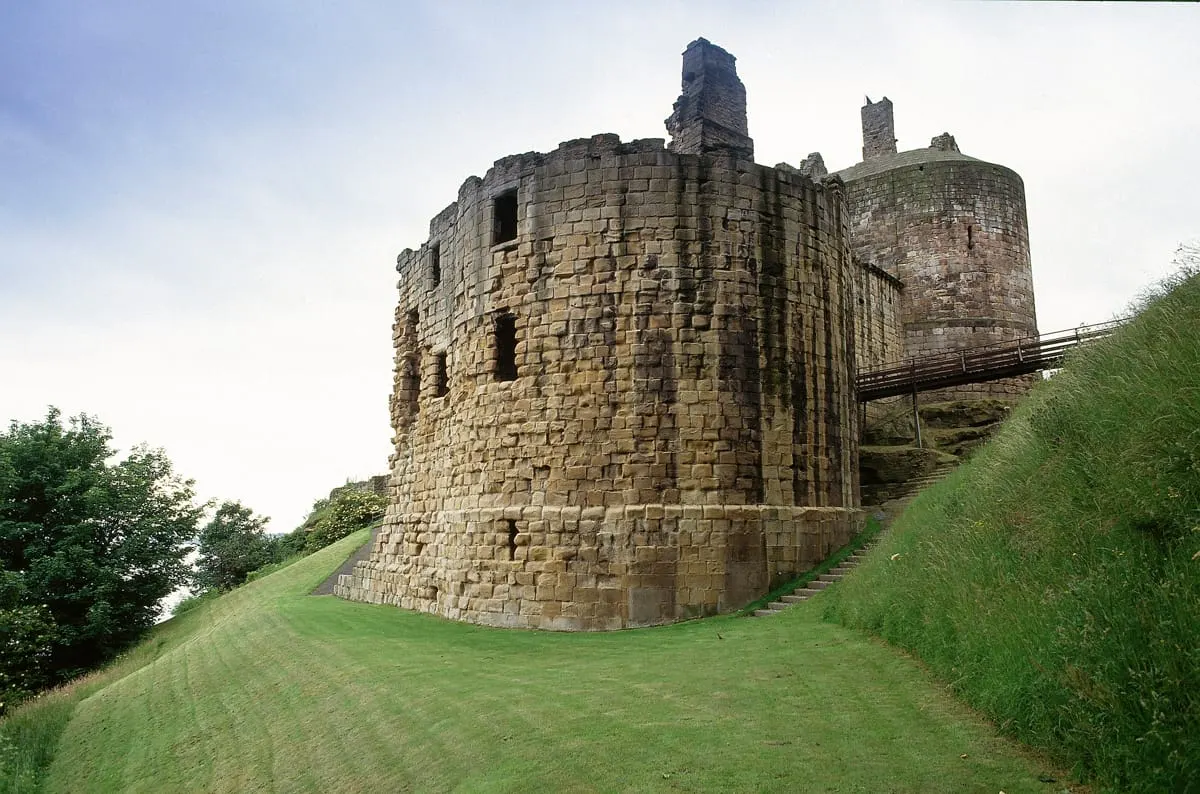 Ruined medieval stone castle with circular towers set on a grassy mound beneath overcast sky.
