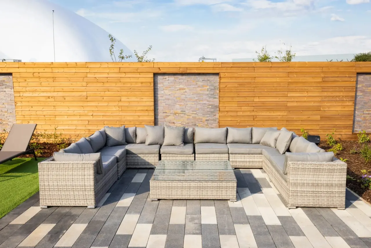 Rattan garden furniture with grey cushions arranged in a U-shape on a patterned patio, backed by wooden fencing.