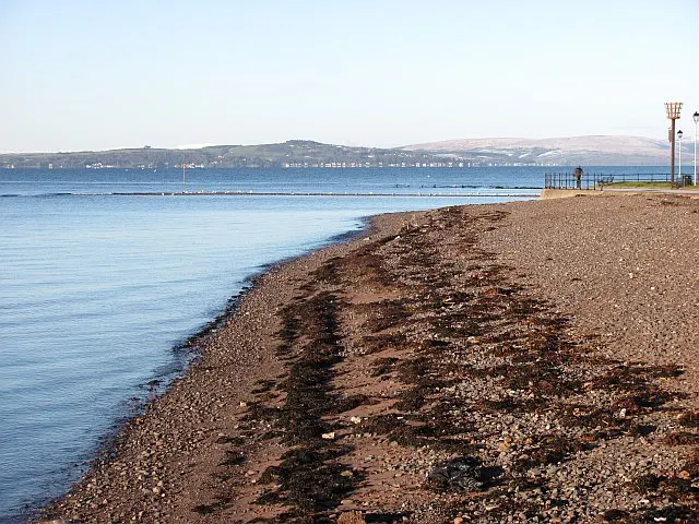 Pebble beach with calm blue water, distant shoreline and hills under clear sky.