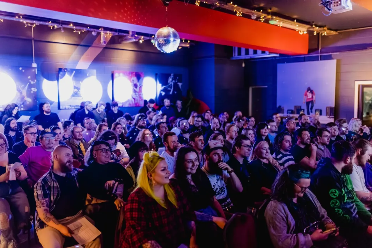Large crowd of people watching a live performance in a colourful indoor venue with stage lighting and a disco ball.
