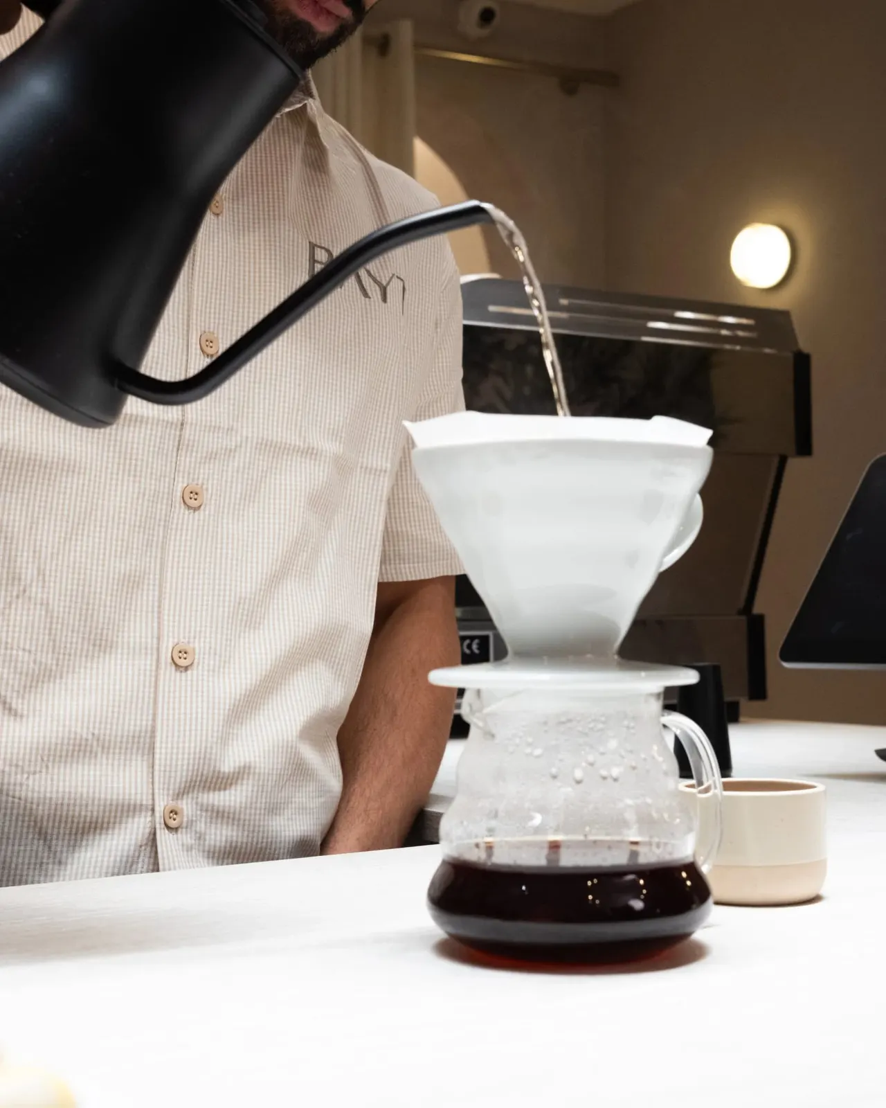 A man in a pale pink shirt pours hot water from a black kettle over a white pour-over coffee filter into a glass carafe.