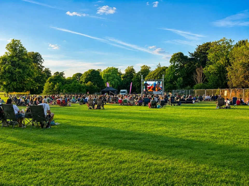 Large outdoor gathering with hundreds of people seated on grass watching a screen, surrounded by trees under blue sky.