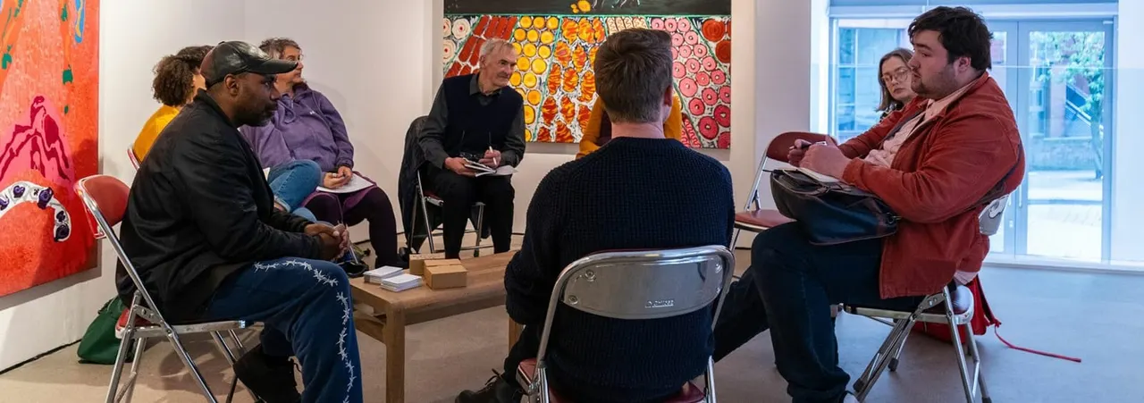 Group of people seated in a circle during an art discussion or workshop in a gallery space with colorful artworks on the walls.
