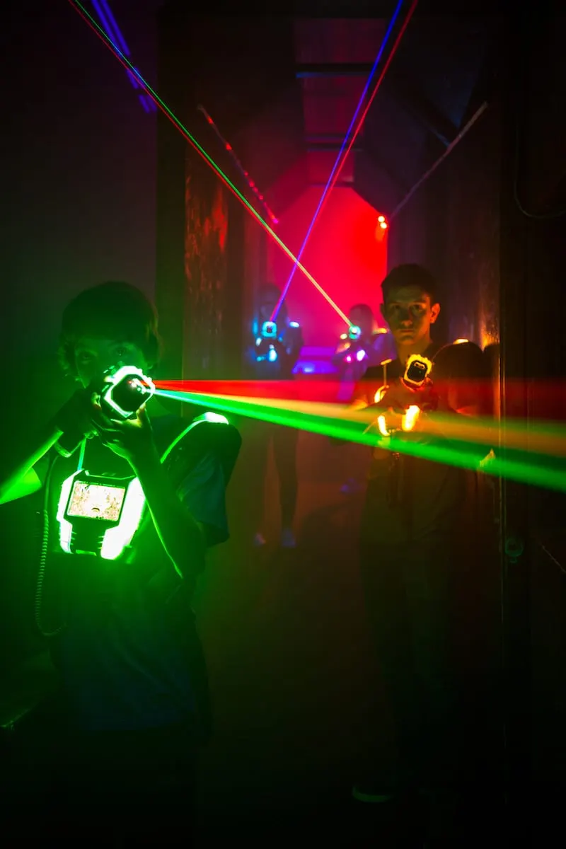 Players in a laser tag arena illuminated by neon green, red and blue laser beams crossing through the space.