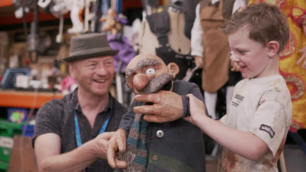 A young boy smiles whilst holding a carved wooden puppet, guided by a puppeteer in a hat at a busy venue.