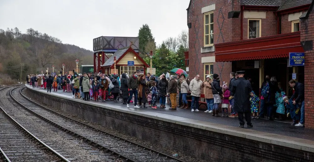 Crowds of visitors and families waiting on a railway platform beside brick station buildings and train tracks.