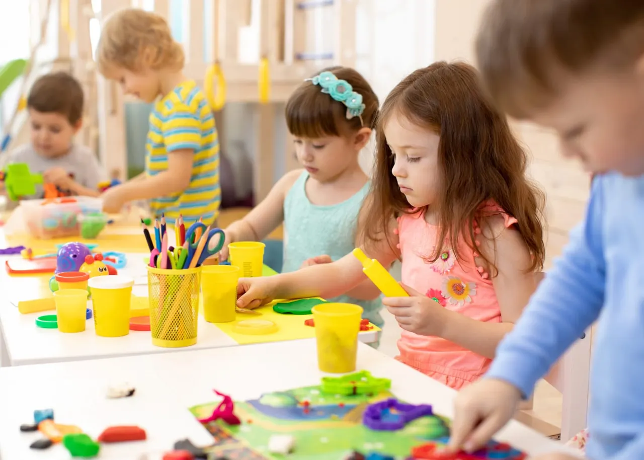 Young children engaged in arts and crafts activities at a table with coloured pencils, paint cups, and craft materials.