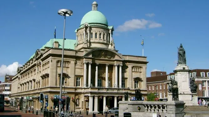 Historic stone building with green dome and classical columns, with statue and pedestrians in town square.