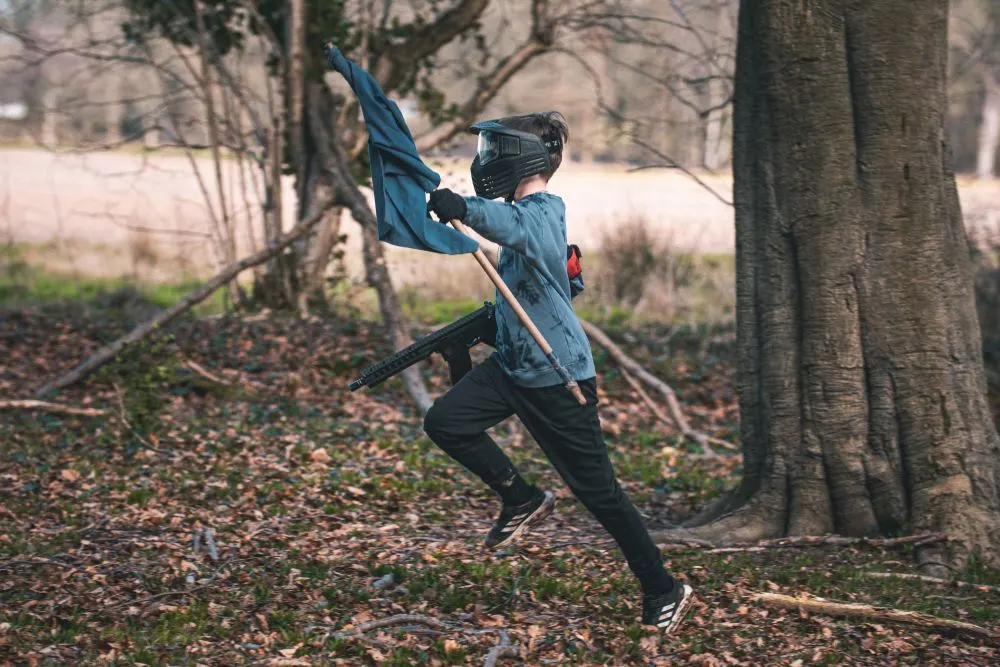 Person in protective gear playing paintball in a woodland area, holding a paintball marker and flag.