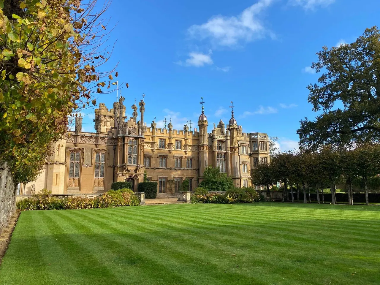 A grand Victorian Gothic stone mansion surrounded by manicured lawns and autumn foliage under clear blue sky.
