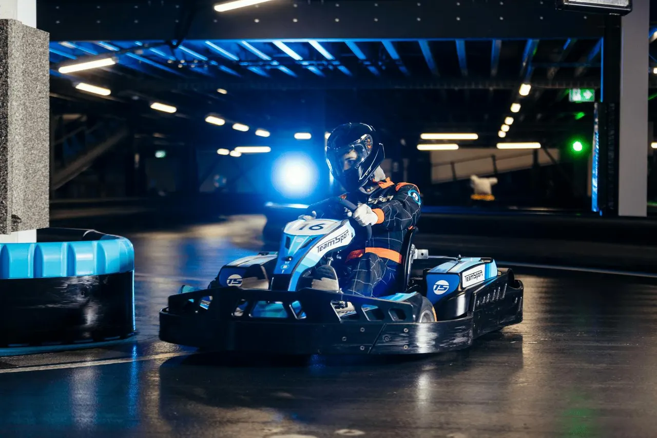 A racer wearing a helmet and protective suit drives a blue and white go-kart around an indoor track lit with blue lighting and barriers.