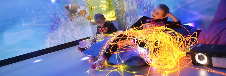 Two young children interact with colourful glowing light installations in an immersive play environment.