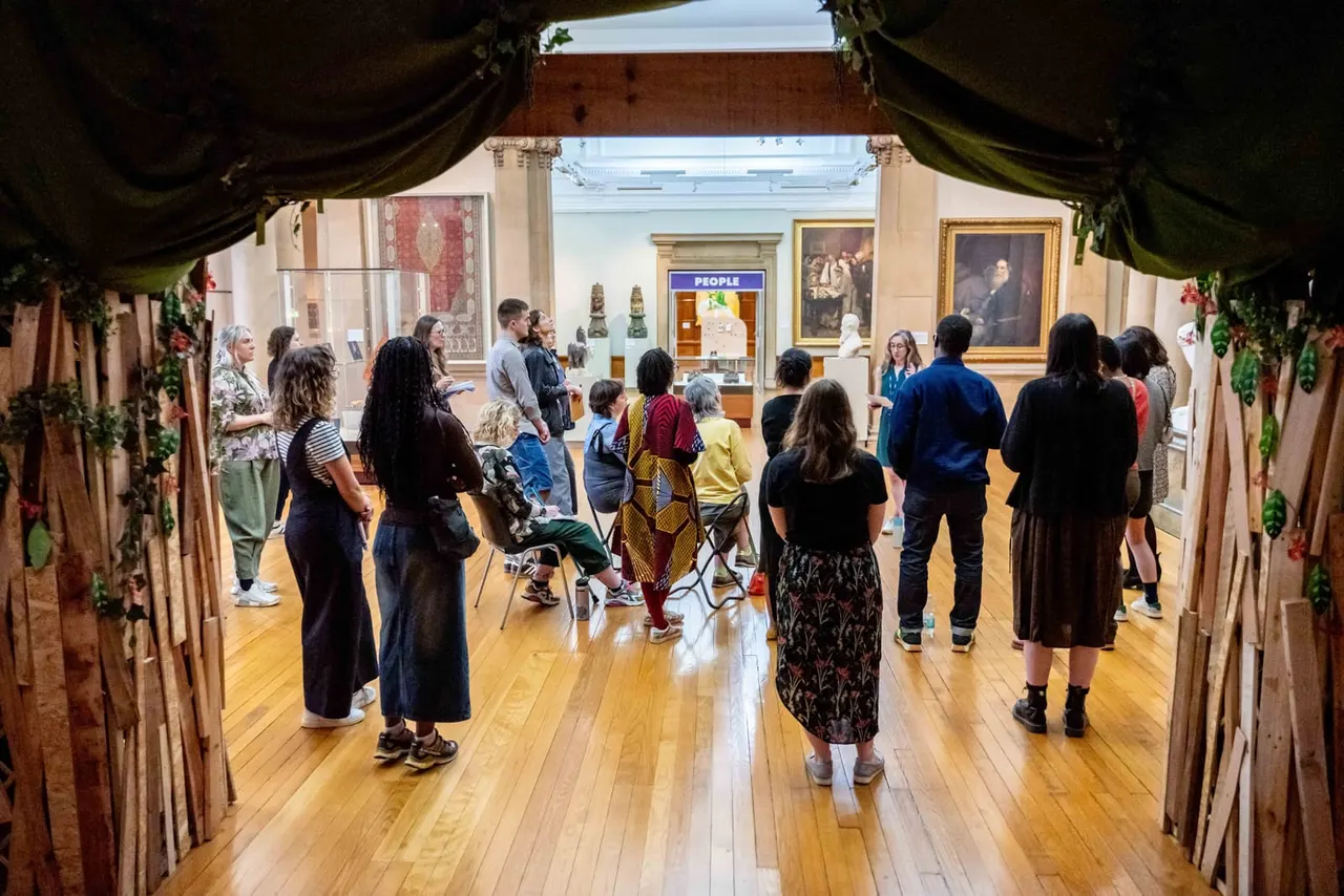 Visitors of mixed ages stand and walk through a museum gallery space with wooden flooring, viewing displays and portraits on the walls.