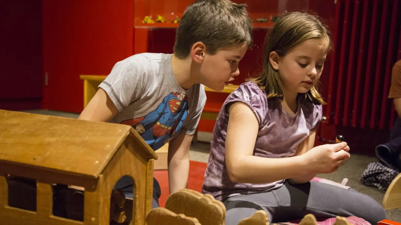 Two children sitting on the floor engaging with wooden play structures in a vibrant red-walled indoor play area.