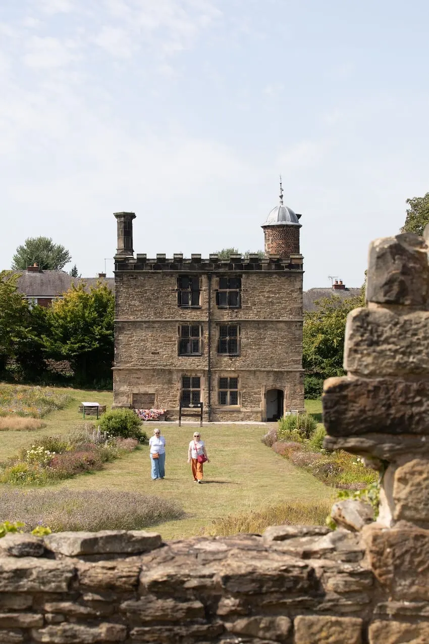Two visitors walk across a grassy courtyard towards a three-storey stone tower house with battlements and a domed turret, surrounded by gardens and ancient stone ruins.
