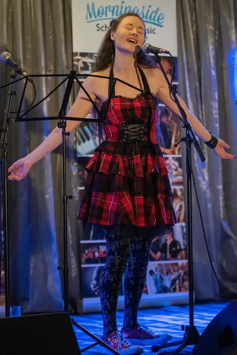 A young woman performs at a microphone on stage, wearing a red tartan dress and patterned tights, singing with expression.
