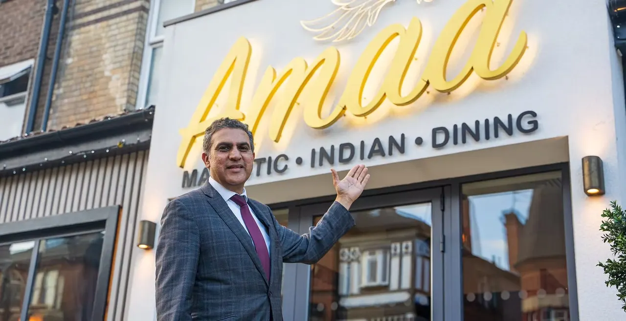 Man in business suit gesturing towards the entrance of an Indian restaurant with a yellow neon sign.