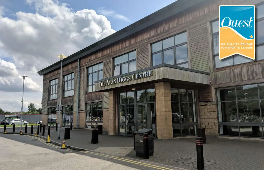Modern leisure centre building with wooden cladding and large windows, displaying Quest quality scheme badge, with car park visible to the left.