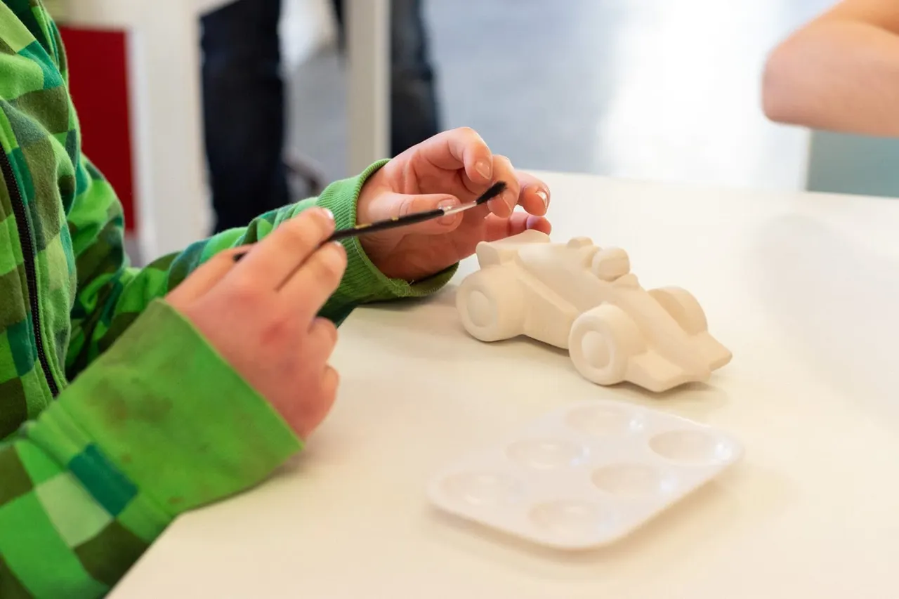 Child in green jacket painting a white ceramic car model at a table.