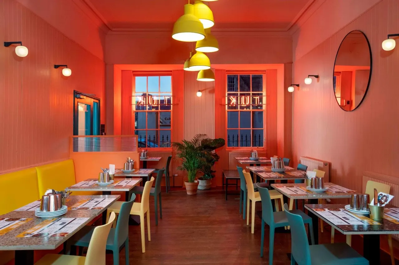 Colourful restaurant interior with bright red walls, yellow pendant lights, dining tables set with place settings, and mixed coloured chairs.