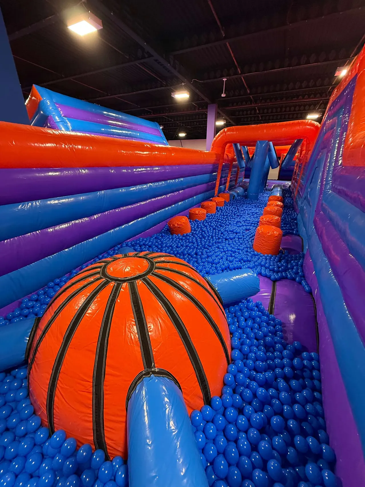 Indoor inflatable play structure with colourful bouncy castles, ball pit, and obstacles in bright blue, red, and purple.