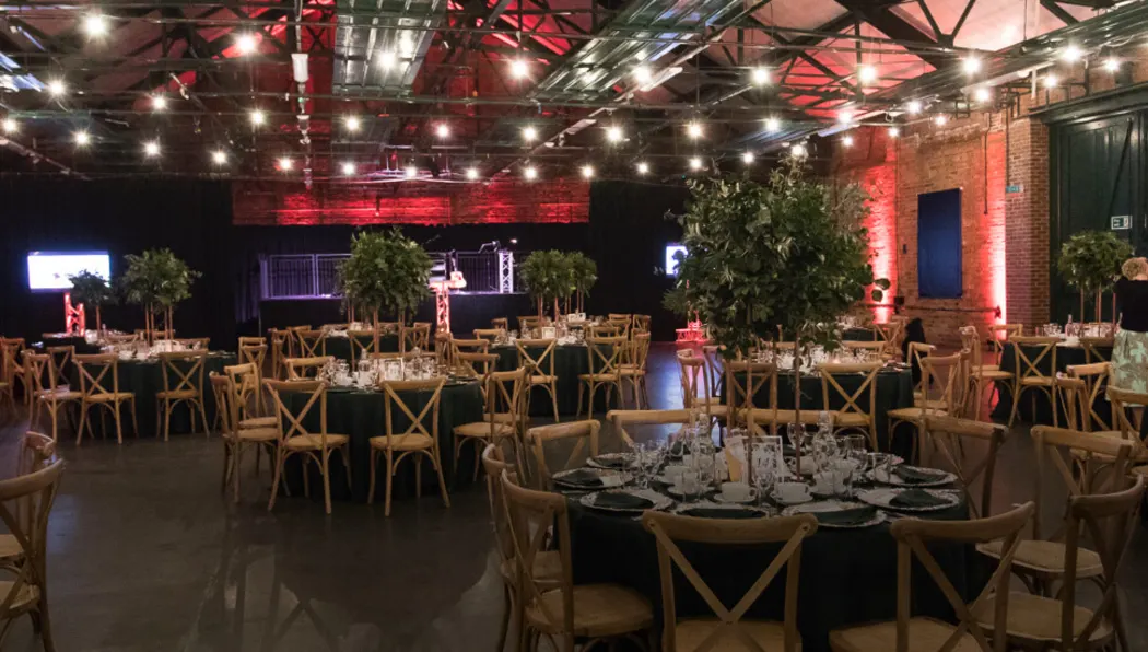 Industrial event space with round tables set for dinner, wooden chairs, red atmospheric lighting and exposed brick walls.