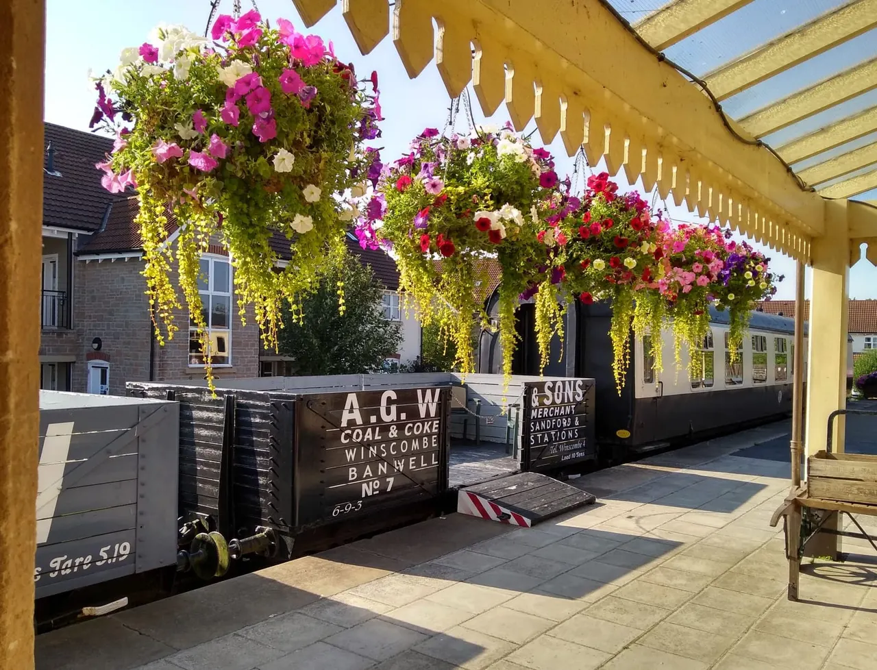 Hanging baskets overflowing with pink, magenta and white flowers suspended from a yellow pergola above a paved courtyard with heritage railway wagons.