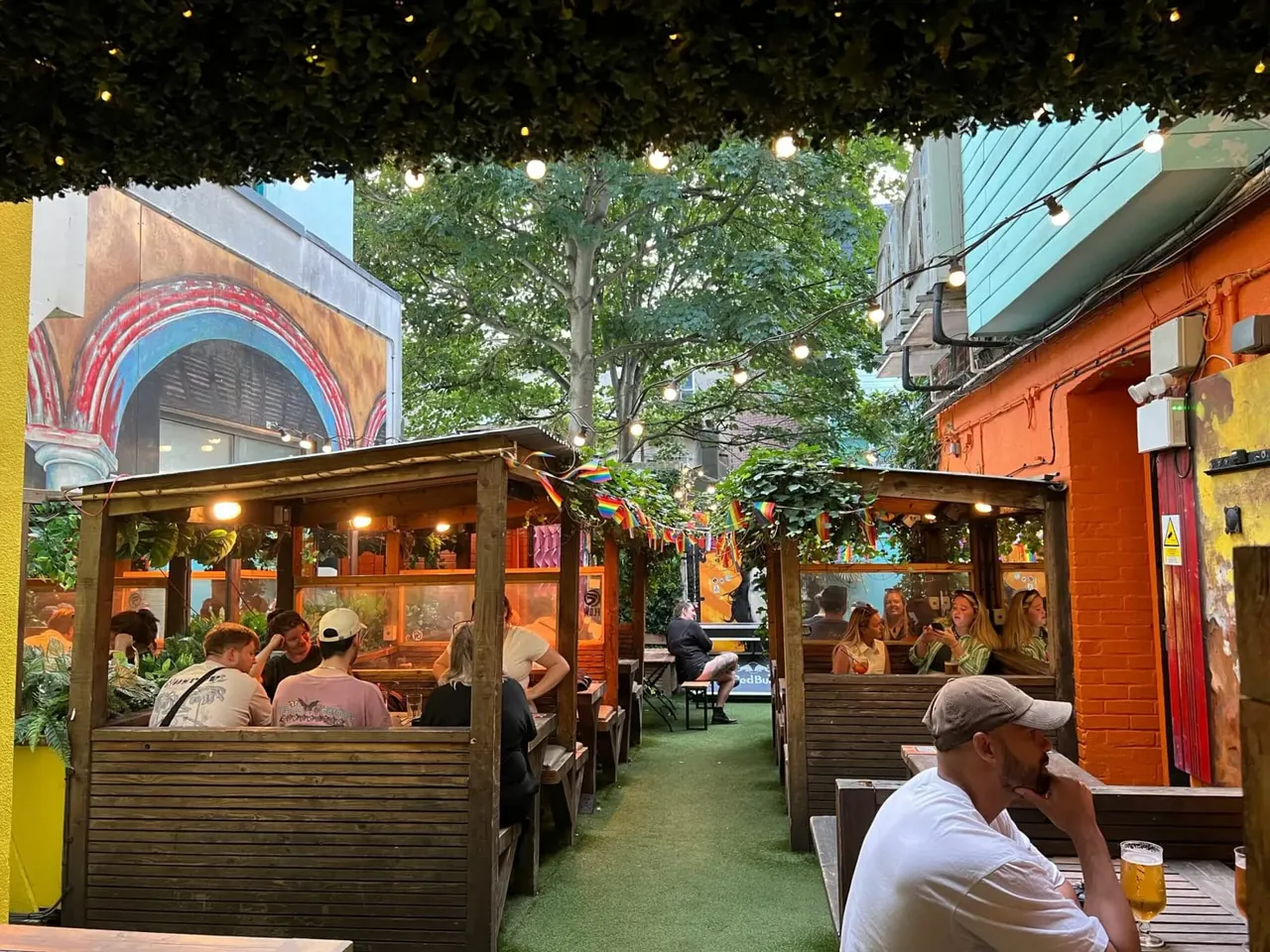 Outdoor beer garden with wooden seating booths, green artificial turf, festoon lights and ivy overhead, colourful buildings surrounding the space, and visitors enjoying drinks.