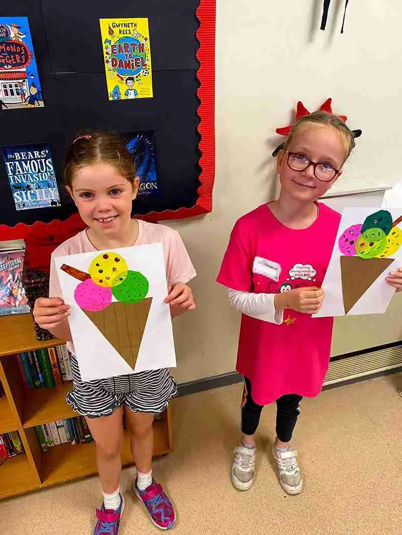 Two young girls display colourful handmade ice cream cone crafts in a classroom decorated with children's book posters.