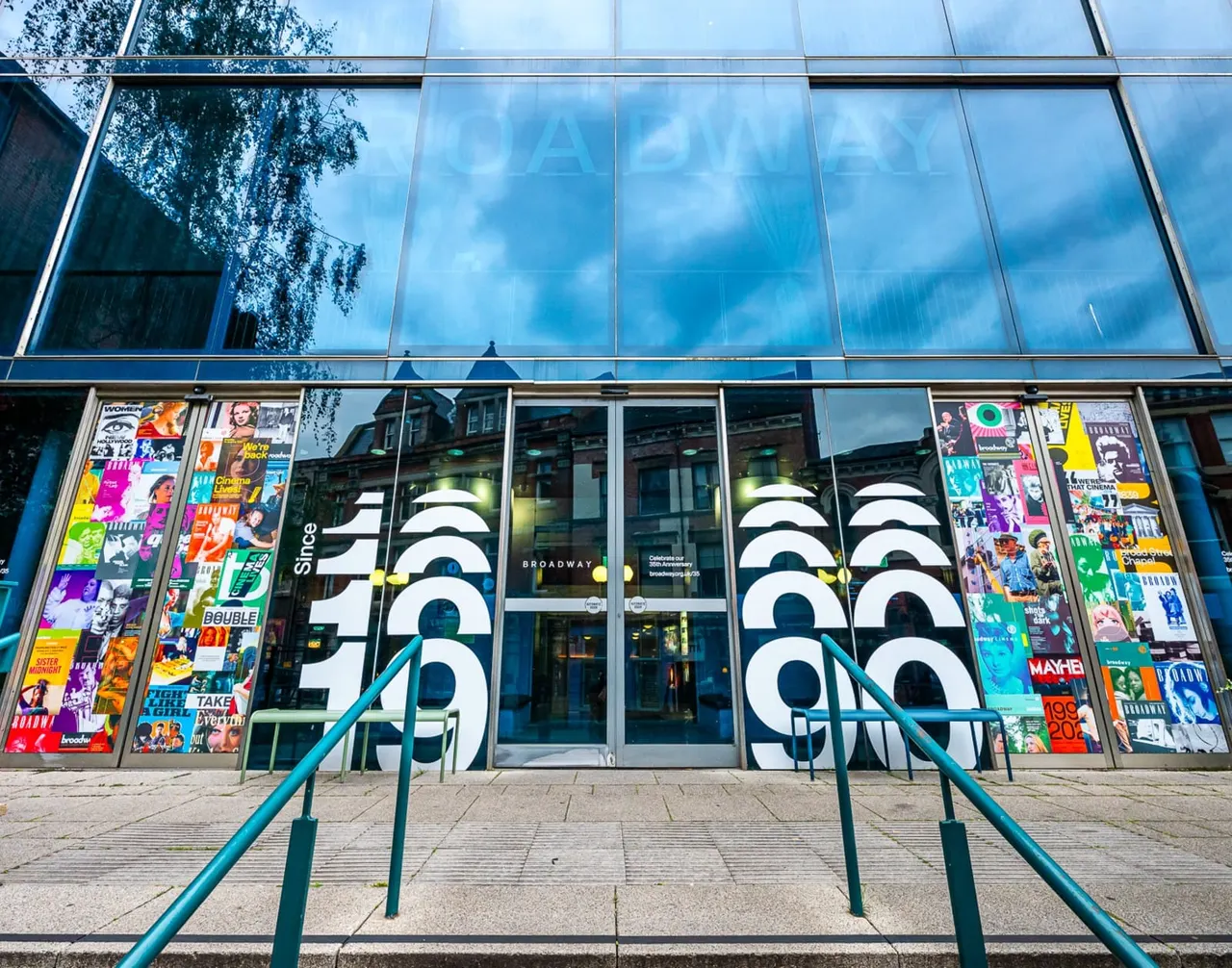 Modern glass-fronted cinema building with colourful posters flanking the entrance doors and blue metal railings leading up to the entrance.