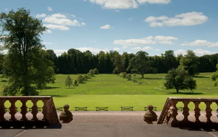 View from a stone balustrade across expansive parkland with scattered trees, green fields and grazing sheep under blue sky.