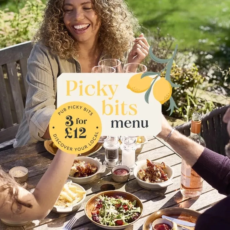 A woman holding a "Picky bits menu" promotional sign smiles at a garden table with food and drinks shared with another person.