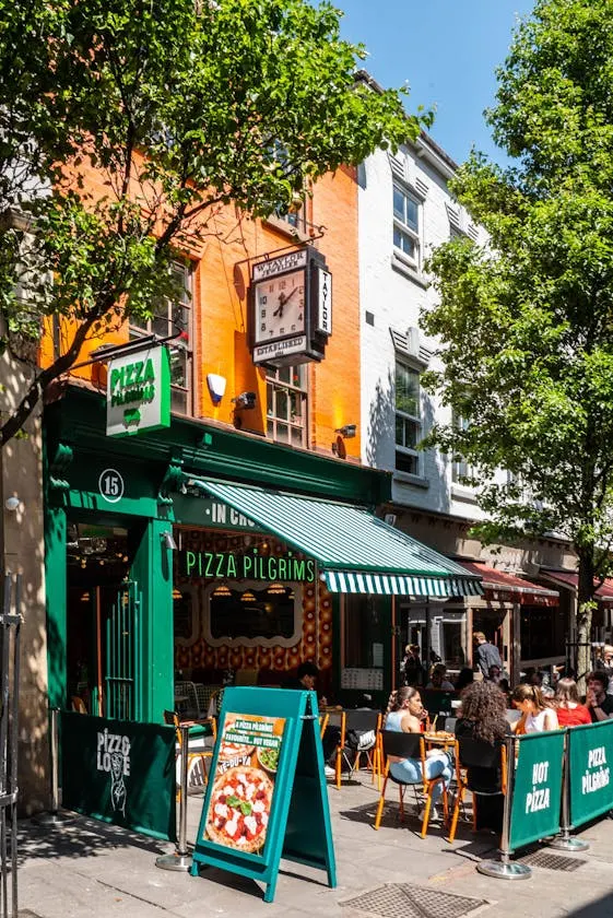 Busy street-level pizza restaurant with green storefront, outdoor seating with customers, trees overhead, and period building facade above.