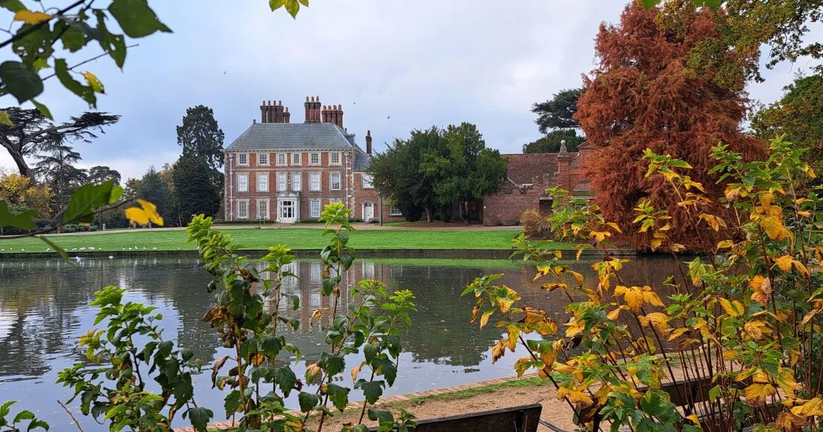 Georgian mansion reflected in a still pond, surrounded by autumn foliage and manicured grounds.