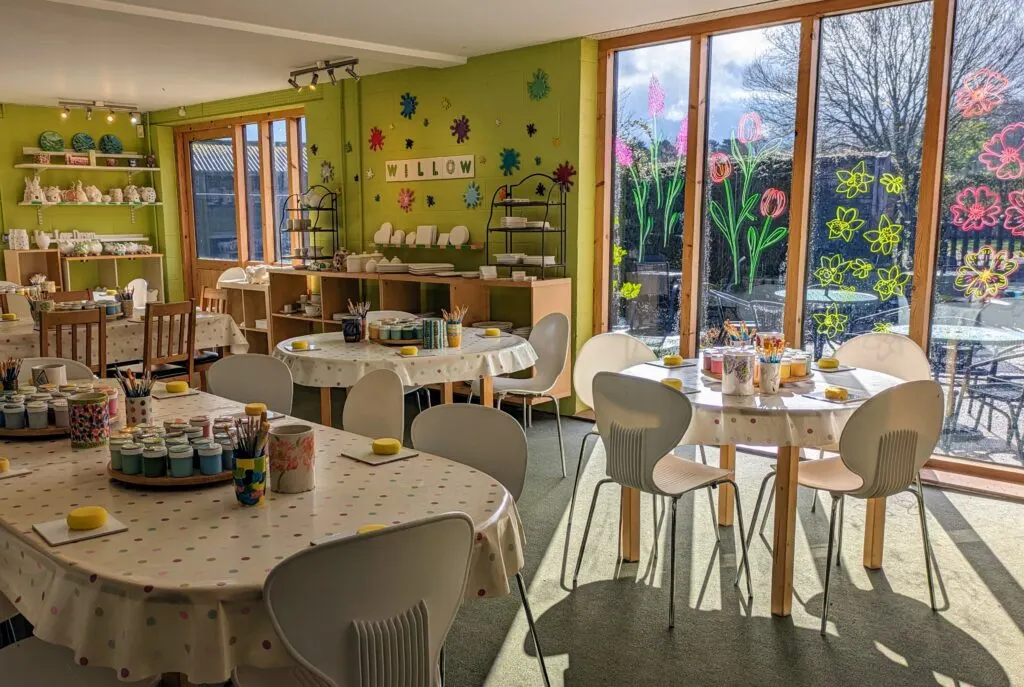 Bright, colourful children's craft studio with multiple tables, chairs, art supplies and large windows displaying flower window paintings.