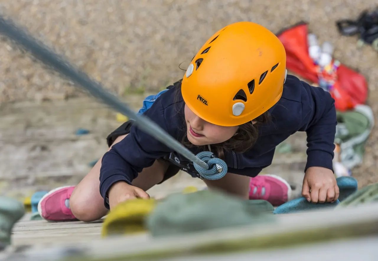 Child in yellow helmet and safety harness climbing an indoor rock wall with coloured hand and foot holds.