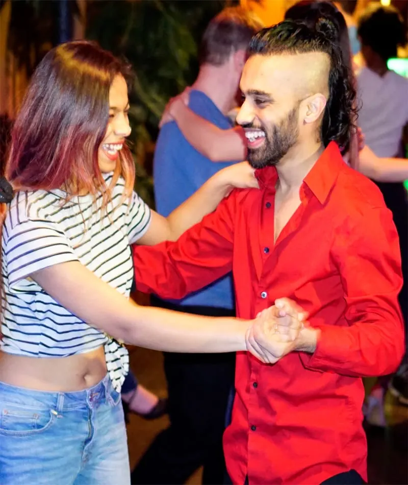 A man in a red shirt and a woman in a striped top dance together at a social dance event with other dancers in the background.
